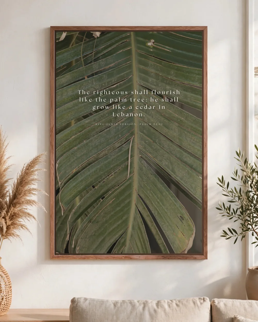 Psalm 92:12 in spaced white serif across a close-up palm frond, pecan frame, hung above a linen sofa.