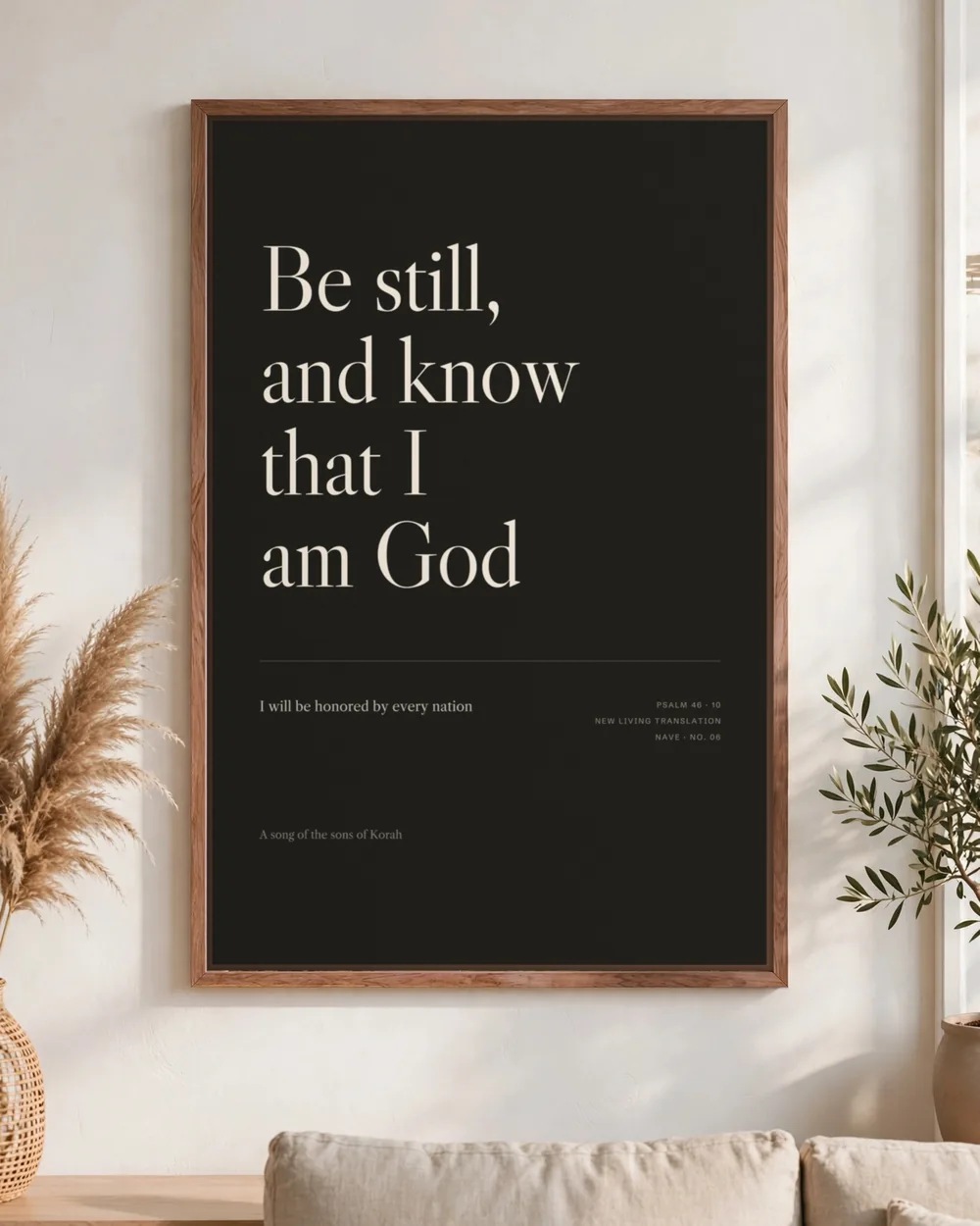 “Be still, and know that I am God” in cream serif on near-black canvas, pecan frame, hung above a linen sofa.