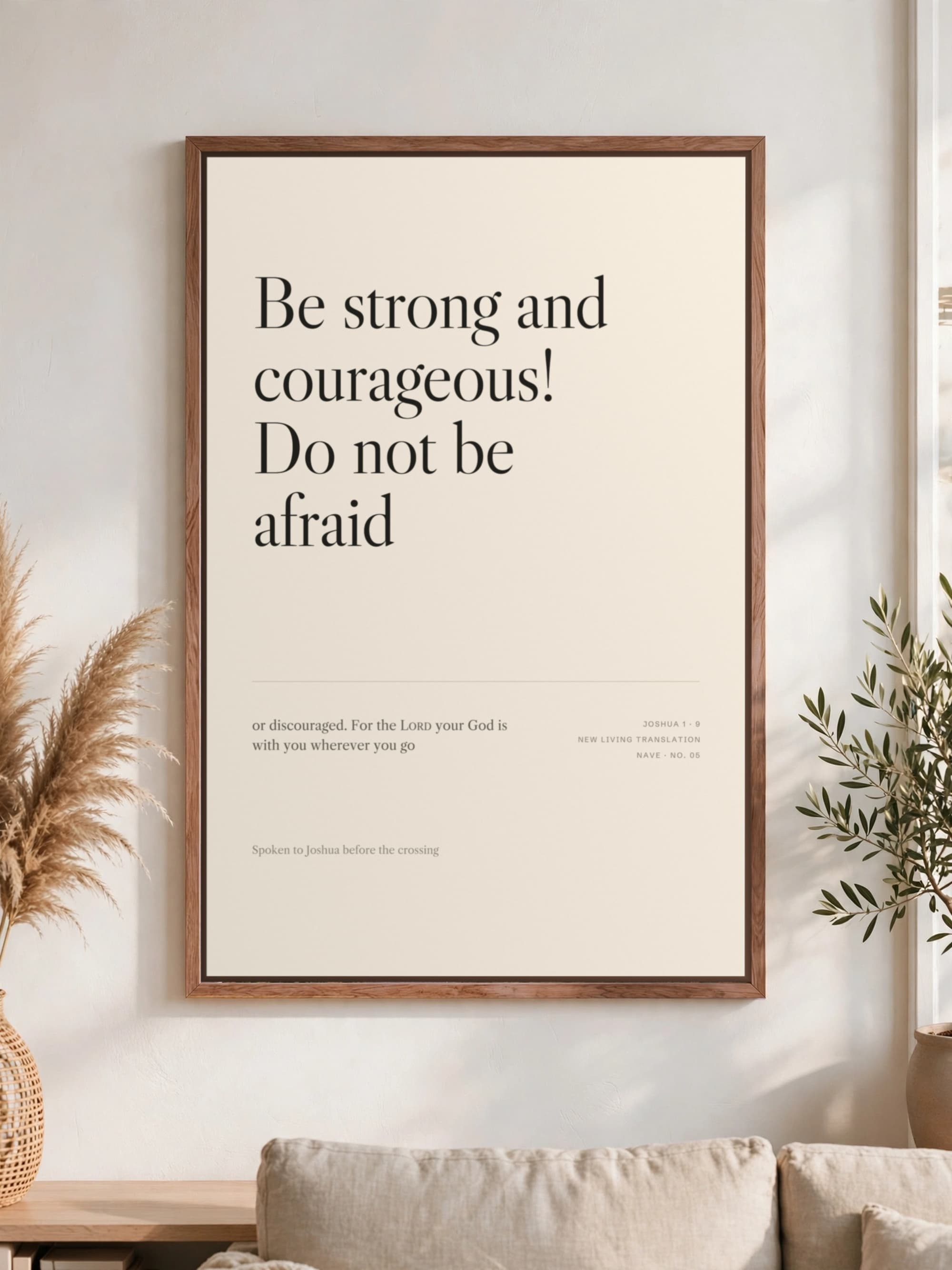“Be strong and courageous” in dark serif on cream canvas, pecan frame, hung above a linen sofa.