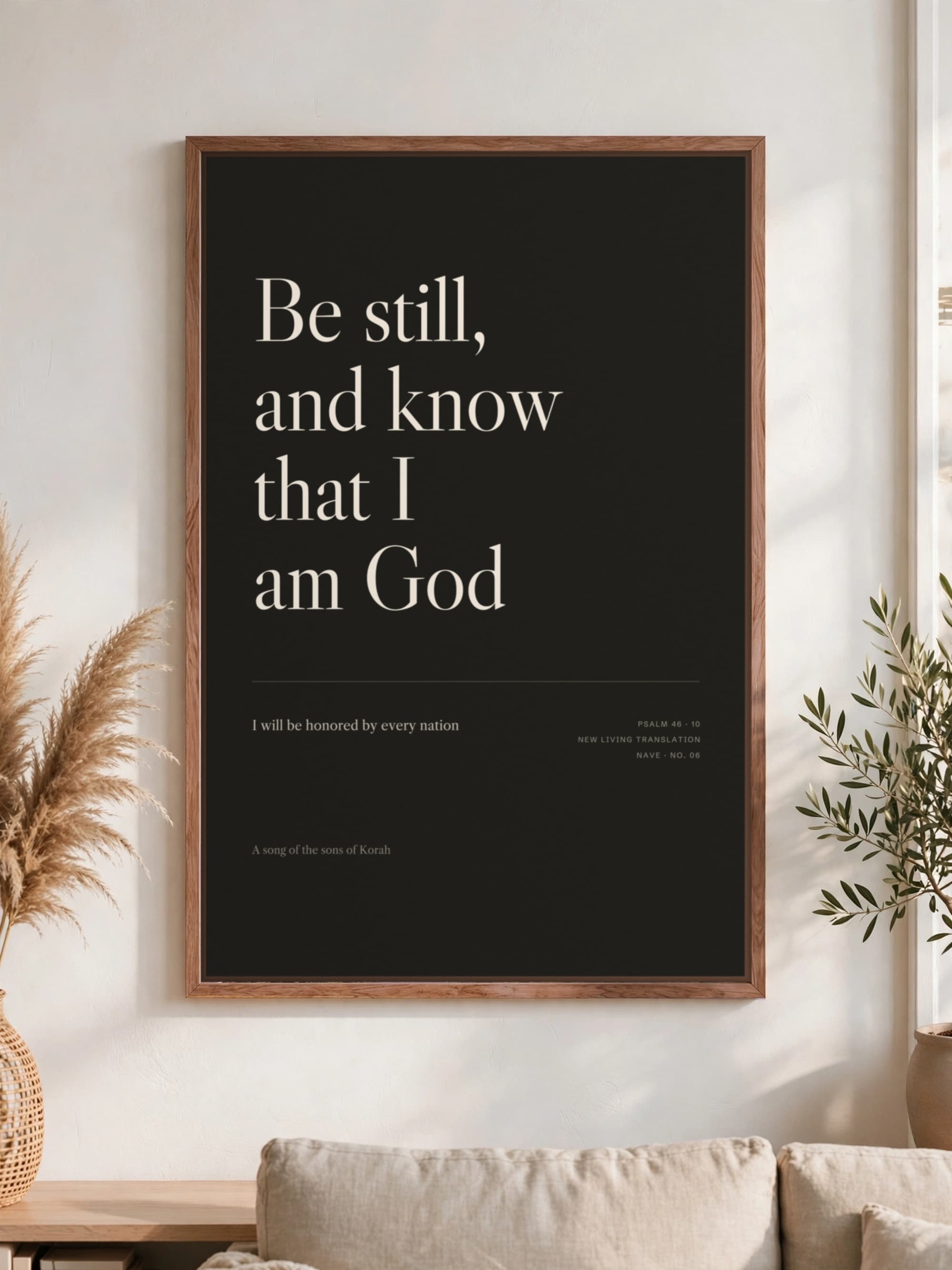 “Be still, and know that I am God” in cream serif on near-black canvas, pecan frame, hung above a linen sofa.