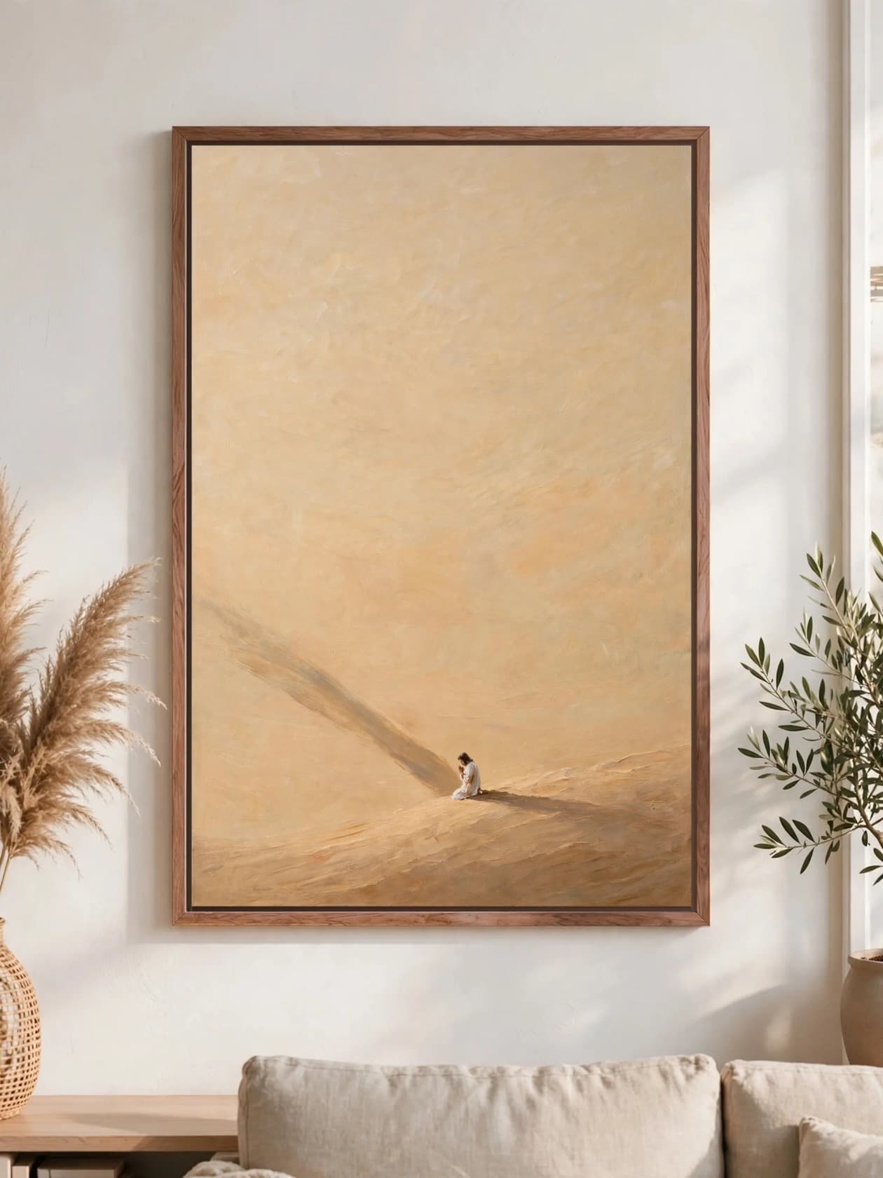 A figure in white kneeling in prayer on a warm sand slope, a long shadow behind, pecan frame, hung above a linen sofa.