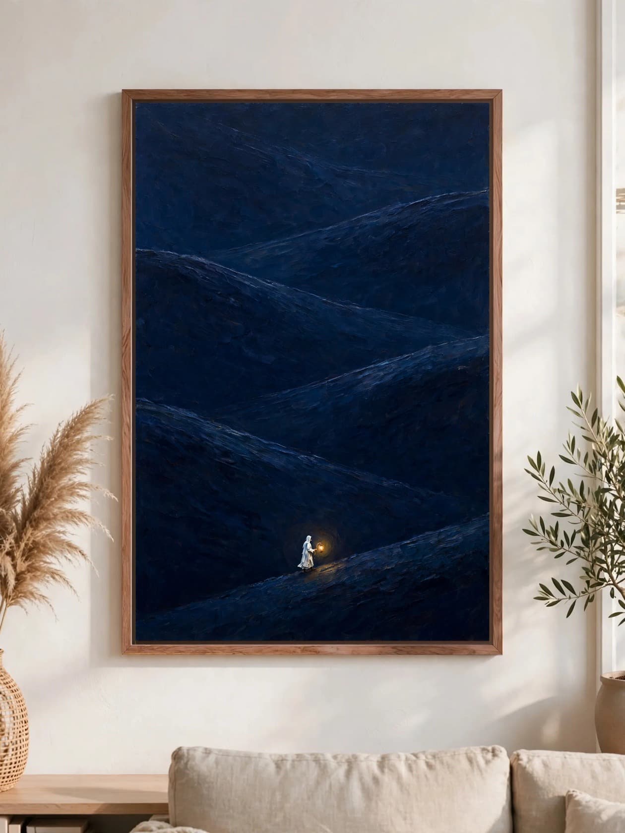 A figure in white carrying a glowing lantern across dark navy hills at night, pecan frame, hung above a linen sofa.